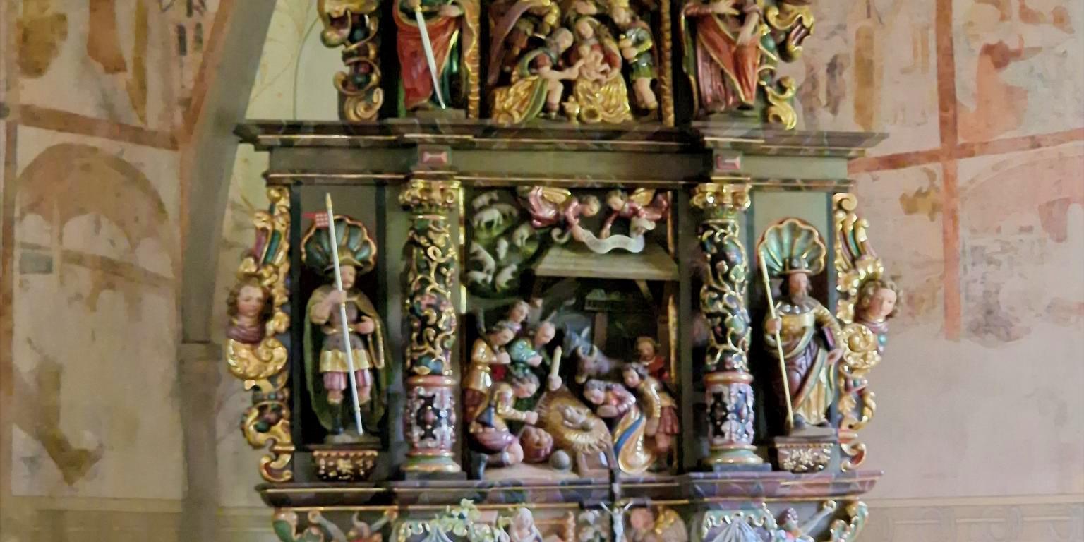 Stellen Sie sich vor, Sie stehen oder sitzen in einer kleinen Kapelle. Vor Ihnen befindet sich ein reich verzierter, farbig gefasster Altar aus Holz. Der Altar ist mehrstöckig aufgebaut und wirkt wie ein kleines, kunstvolles Haus mit Säulen, Figuren und Nischen. Die Farben sind warm: Gold, Rot, Grün und dunkles Holz prägen das Gesamtbild. Im Zentrum des Altars, auf Augenhöhe, sehen Sie eine Darstellung der Geburt Jesu: Ein kleines Kind liegt in der Mitte, umgeben von mehreren Figuren. Einige Figuren knien, andere stehen. Alle sind dicht um die Mitte gruppiert, sodass ein Gefühl von Nähe und Sammlung entsteht. Darüber, auf einer erhöhten Ebene, befinden sich weitere Figuren, die Szenen aus dem Leben Jesu oder aus der biblischen Geschichte zeigen. Diese Figuren sind kleiner, aber ebenfalls reich gestaltet. Auch hier stehen mehrere Gestalten beieinander, eingerahmt von Säulen und Bögen. Seitlich stehen größere Figuren in Nischen. Jede Figur ist einzeln hervorgehoben, mit Gewändern in kräftigen Farben und goldenen Details. Unterhalb der Hauptszene gibt es eine weitere kleine Darstellung, ebenfalls mit mehreren Figuren, die sich um eine zentrale Szene gruppieren. Vor dem Altar steht ein schlichter, heller Altartisch mit einem weißen Tuch. Darauf befinden sich frische Blumen. Im Hintergrund erkennt man die Wände der Kapelle mit zarten, teilweise verblassten Malereien. Die Decke ist gewölbt und trägt ebenfalls helle, zurückhaltende Bildmotive. Insgesamt entsteht ein Eindruck von Stille, Konzentration und Tiefe: Der kunstreiche Altar lädt dazu ein, den Blick immer wieder zur Mitte zu führen – dorthin, wo das Wesentliche liegt.