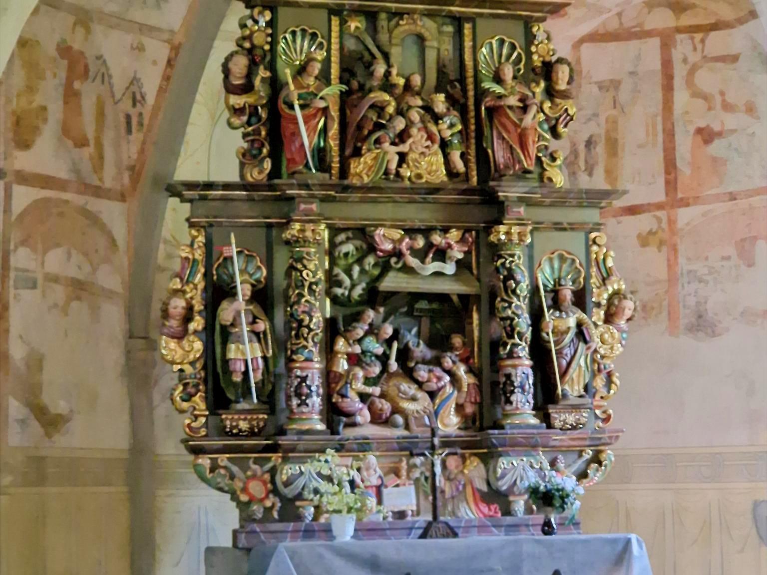 Stellen Sie sich vor, Sie stehen oder sitzen in einer kleinen Kapelle. Vor Ihnen befindet sich ein reich verzierter, farbig gefasster Altar aus Holz. Der Altar ist mehrstöckig aufgebaut und wirkt wie ein kleines, kunstvolles Haus mit Säulen, Figuren und Nischen. Die Farben sind warm: Gold, Rot, Grün und dunkles Holz prägen das Gesamtbild. Im Zentrum des Altars, auf Augenhöhe, sehen Sie eine Darstellung der Geburt Jesu: Ein kleines Kind liegt in der Mitte, umgeben von mehreren Figuren. Einige Figuren knien, andere stehen. Alle sind dicht um die Mitte gruppiert, sodass ein Gefühl von Nähe und Sammlung entsteht. Darüber, auf einer erhöhten Ebene, befinden sich weitere Figuren, die Szenen aus dem Leben Jesu oder aus der biblischen Geschichte zeigen. Diese Figuren sind kleiner, aber ebenfalls reich gestaltet. Auch hier stehen mehrere Gestalten beieinander, eingerahmt von Säulen und Bögen. Seitlich stehen größere Figuren in Nischen. Jede Figur ist einzeln hervorgehoben, mit Gewändern in kräftigen Farben und goldenen Details. Unterhalb der Hauptszene gibt es eine weitere kleine Darstellung, ebenfalls mit mehreren Figuren, die sich um eine zentrale Szene gruppieren. Vor dem Altar steht ein schlichter, heller Altartisch mit einem weißen Tuch. Darauf befinden sich frische Blumen. Im Hintergrund erkennt man die Wände der Kapelle mit zarten, teilweise verblassten Malereien. Die Decke ist gewölbt und trägt ebenfalls helle, zurückhaltende Bildmotive. Insgesamt entsteht ein Eindruck von Stille, Konzentration und Tiefe: Der kunstreiche Altar lädt dazu ein, den Blick immer wieder zur Mitte zu führen – dorthin, wo das Wesentliche liegt.