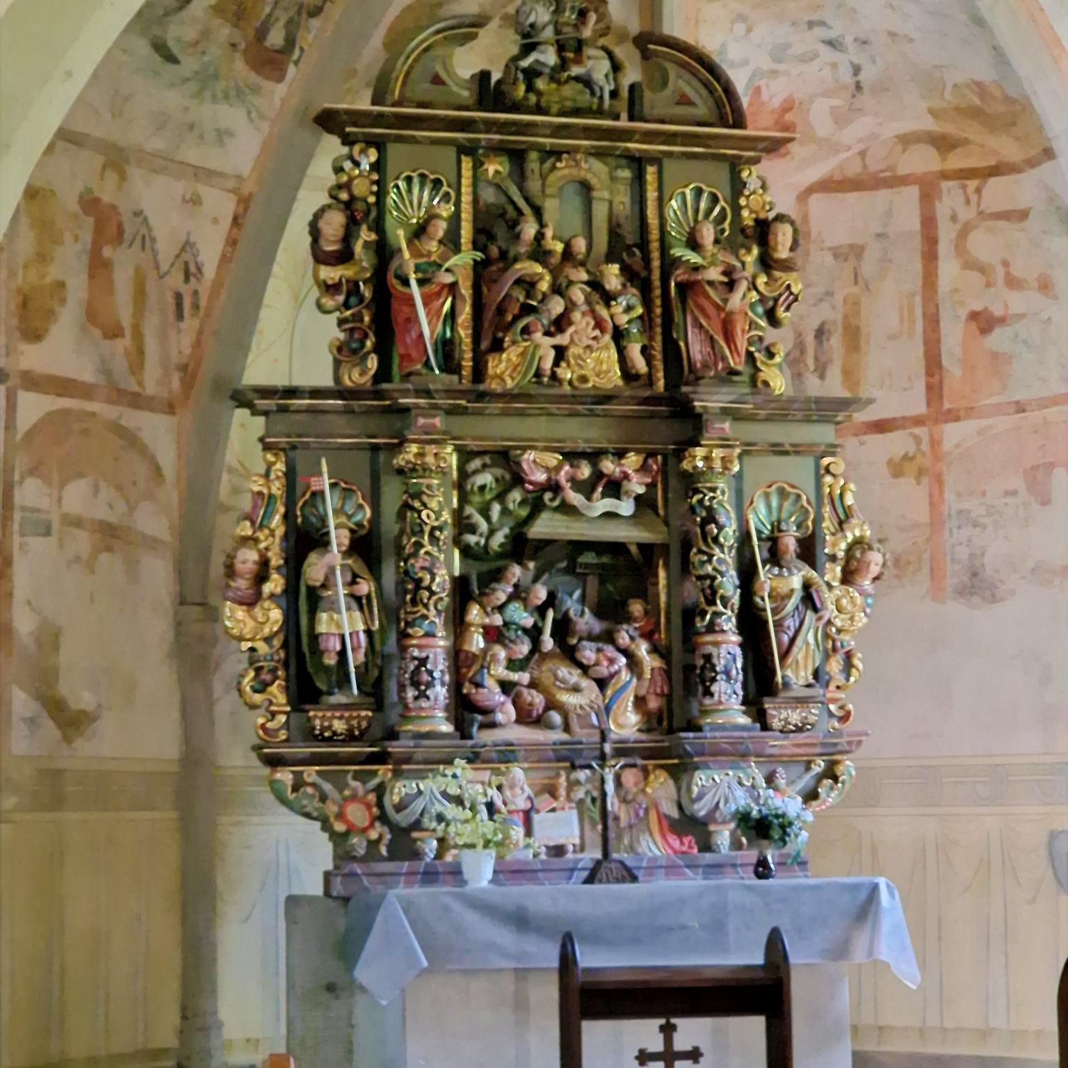 Stellen Sie sich vor, Sie stehen oder sitzen in einer kleinen Kapelle. Vor Ihnen befindet sich ein reich verzierter, farbig gefasster Altar aus Holz. Der Altar ist mehrstöckig aufgebaut und wirkt wie ein kleines, kunstvolles Haus mit Säulen, Figuren und Nischen. Die Farben sind warm: Gold, Rot, Grün und dunkles Holz prägen das Gesamtbild. Im Zentrum des Altars, auf Augenhöhe, sehen Sie eine Darstellung der Geburt Jesu: Ein kleines Kind liegt in der Mitte, umgeben von mehreren Figuren. Einige Figuren knien, andere stehen. Alle sind dicht um die Mitte gruppiert, sodass ein Gefühl von Nähe und Sammlung entsteht. Darüber, auf einer erhöhten Ebene, befinden sich weitere Figuren, die Szenen aus dem Leben Jesu oder aus der biblischen Geschichte zeigen. Diese Figuren sind kleiner, aber ebenfalls reich gestaltet. Auch hier stehen mehrere Gestalten beieinander, eingerahmt von Säulen und Bögen. Seitlich stehen größere Figuren in Nischen. Jede Figur ist einzeln hervorgehoben, mit Gewändern in kräftigen Farben und goldenen Details. Unterhalb der Hauptszene gibt es eine weitere kleine Darstellung, ebenfalls mit mehreren Figuren, die sich um eine zentrale Szene gruppieren. Vor dem Altar steht ein schlichter, heller Altartisch mit einem weißen Tuch. Darauf befinden sich frische Blumen. Im Hintergrund erkennt man die Wände der Kapelle mit zarten, teilweise verblassten Malereien. Die Decke ist gewölbt und trägt ebenfalls helle, zurückhaltende Bildmotive. Insgesamt entsteht ein Eindruck von Stille, Konzentration und Tiefe: Der kunstreiche Altar lädt dazu ein, den Blick immer wieder zur Mitte zu führen – dorthin, wo das Wesentliche liegt.