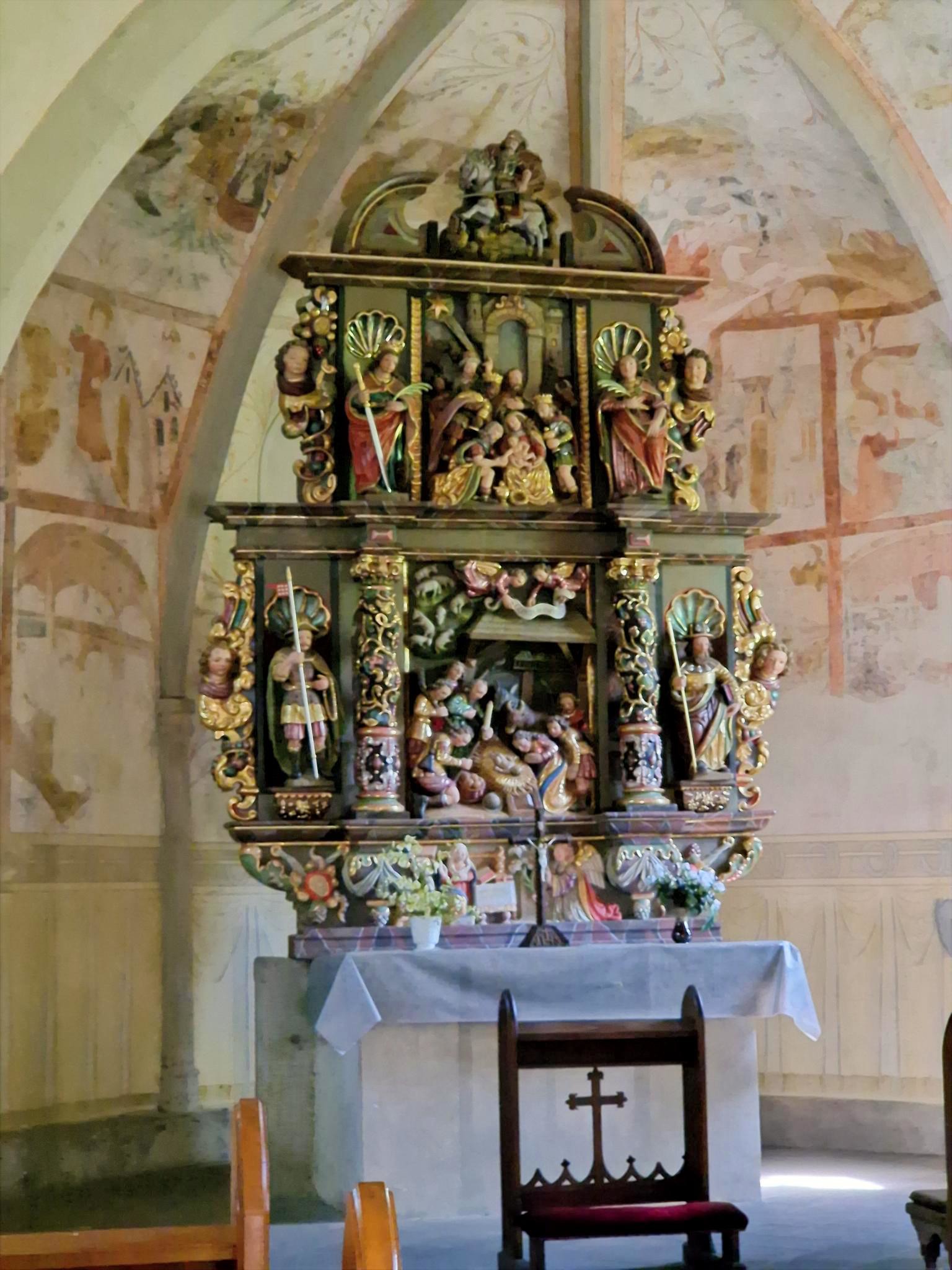 Stellen Sie sich vor, Sie stehen oder sitzen in einer kleinen Kapelle. Vor Ihnen befindet sich ein reich verzierter, farbig gefasster Altar aus Holz. Der Altar ist mehrstöckig aufgebaut und wirkt wie ein kleines, kunstvolles Haus mit Säulen, Figuren und Nischen. Die Farben sind warm: Gold, Rot, Grün und dunkles Holz prägen das Gesamtbild. Im Zentrum des Altars, auf Augenhöhe, sehen Sie eine Darstellung der Geburt Jesu: Ein kleines Kind liegt in der Mitte, umgeben von mehreren Figuren. Einige Figuren knien, andere stehen. Alle sind dicht um die Mitte gruppiert, sodass ein Gefühl von Nähe und Sammlung entsteht. Darüber, auf einer erhöhten Ebene, befinden sich weitere Figuren, die Szenen aus dem Leben Jesu oder aus der biblischen Geschichte zeigen. Diese Figuren sind kleiner, aber ebenfalls reich gestaltet. Auch hier stehen mehrere Gestalten beieinander, eingerahmt von Säulen und Bögen. Seitlich stehen größere Figuren in Nischen. Jede Figur ist einzeln hervorgehoben, mit Gewändern in kräftigen Farben und goldenen Details. Unterhalb der Hauptszene gibt es eine weitere kleine Darstellung, ebenfalls mit mehreren Figuren, die sich um eine zentrale Szene gruppieren. Vor dem Altar steht ein schlichter, heller Altartisch mit einem weißen Tuch. Darauf befinden sich frische Blumen. Im Hintergrund erkennt man die Wände der Kapelle mit zarten, teilweise verblassten Malereien. Die Decke ist gewölbt und trägt ebenfalls helle, zurückhaltende Bildmotive. Insgesamt entsteht ein Eindruck von Stille, Konzentration und Tiefe: Der kunstreiche Altar lädt dazu ein, den Blick immer wieder zur Mitte zu führen – dorthin, wo das Wesentliche liegt.