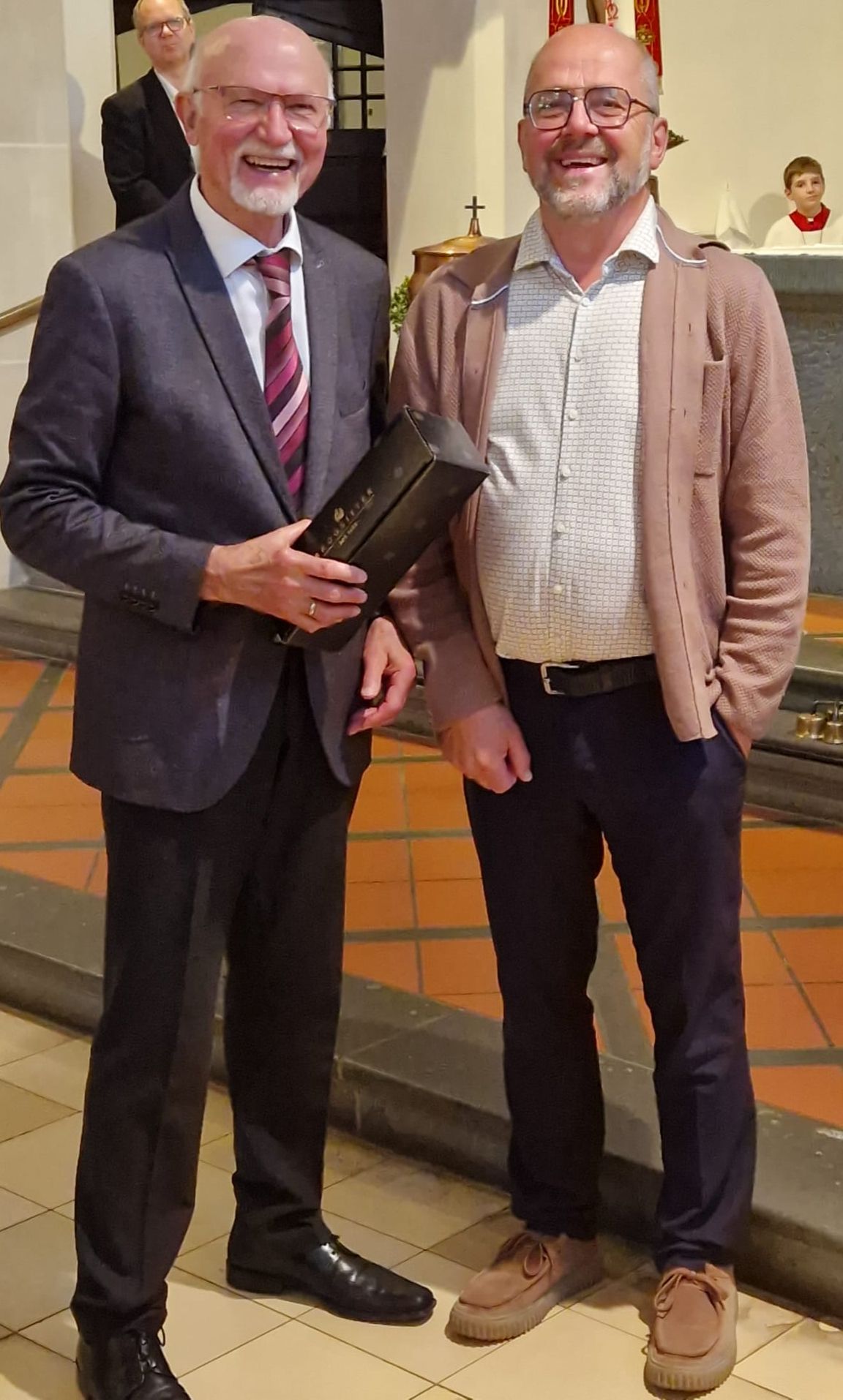Das Foto zeigt zwei Personen, die nebeneinander in einer Kirche stehen. Beide schauen in Richtung Kamera. Die Person links trägt einen dunklen Anzug mit Hemd und gestreifter Krawatte und hält eine rechteckige, schwarze Geschenkbox in der Hand. Die Person rechts trägt eine helle Hose, ein gemustertes Hemd und darüber eine hellbraune Strickjacke. Der Boden besteht aus Fliesen und leicht erhöhten Stufen. Im Hintergrund ist der Altarbereich der Kirche zu sehen, mit liturgischen Gegenständen und weiteren Personen, die unscharf im Hintergrund stehen. Die Szene wirkt wie ein Moment einer Ehrung oder Geschenkübergabe im Rahmen eines Gottesdienstes oder einer Feie (c) Walter Merten Das Foto zeigt zwei Personen, die nebeneinander in einer Kirche stehen. Beide schauen in Richtung Kamera. Die Person links trägt einen dunklen Anzug mit Hemd und gestreifter Krawatte und hält eine rechteckige, schwarze Geschenkbox in der Hand. Die Person rechts trägt eine helle Hose, ein gemustertes Hemd und darüber eine hellbraune Strickjacke. Der Boden besteht aus Fliesen und leicht erhöhten Stufen. Im Hintergrund ist der Altarbereich der Kirche zu sehen, mit liturgischen Gegenständen und weiteren Personen, die unscharf im Hintergrund stehen. Die Szene wirkt wie ein Moment einer Ehrung oder Geschenkübergabe im Rahmen eines Gottesdienstes oder einer Feie