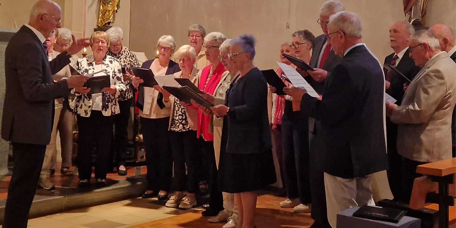 Das Foto zeigt den Kirchenchor Cäcilia Wanderath als Gruppe von etwa 15 bis 20 Personen in der Kirche. Die Gruppe steht in einem Halbkreis und hält Notenmappen in den Händen. Links im Bild steht der Chorleiter Klaus Bierbrauer, die die Gruppe dirigiert und mit erhobener Hand den Takt vorgibt. Die übrigen Personen stehen dicht beieinander auf einer leicht erhöhten Stufe und schauen in ihre Noten oder in Richtung der dirigierenden Person. Die Kleidung ist überwiegend festlich, vor allem dunkle Jacken, mit einigen helleren Oberteilen und farbigen Akzenten. Im Hintergrund ist der Innenraum einer Kirche zu sehen, mit hellen Wänden und einer Figur an der Wand. Der Boden besteht aus hellen Fliesen und Holz. Insgesamt zeigt das Bild eine Chorgemeinschaft. (c) Walter Merten Das Foto zeigt den Kirchenchor Cäcilia Wanderath als Gruppe von etwa 15 bis 20 Personen in der Kirche. Die Gruppe steht in einem Halbkreis und hält Notenmappen in den Händen. Links im Bild steht der Chorleiter Klaus Bierbrauer, die die Gruppe dirigiert und mit erhobener Hand den Takt vorgibt. Die übrigen Personen stehen dicht beieinander auf einer leicht erhöhten Stufe und schauen in ihre Noten oder in Richtung der dirigierenden Person. Die Kleidung ist überwiegend festlich, vor allem dunkle Jacken, mit einigen helleren Oberteilen und farbigen Akzenten. Im Hintergrund ist der Innenraum einer Kirche zu sehen, mit hellen Wänden und einer Figur an der Wand. Der Boden besteht aus hellen Fliesen und Holz. Insgesamt zeigt das Bild eine Chorgemeinschaft.