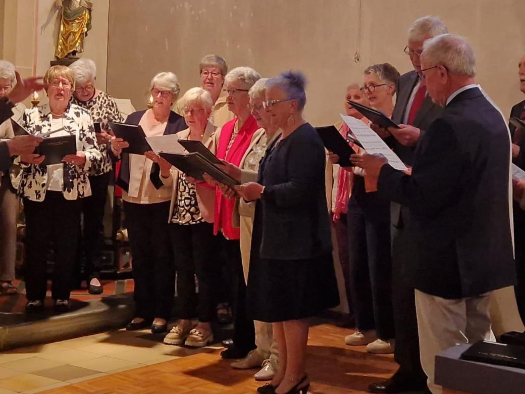 Das Foto zeigt den Kirchenchor Cäcilia Wanderath als Gruppe von etwa 15 bis 20 Personen in der Kirche. Die Gruppe steht in einem Halbkreis und hält Notenmappen in den Händen. Links im Bild steht der Chorleiter Klaus Bierbrauer, die die Gruppe dirigiert und mit erhobener Hand den Takt vorgibt. Die übrigen Personen stehen dicht beieinander auf einer leicht erhöhten Stufe und schauen in ihre Noten oder in Richtung der dirigierenden Person. Die Kleidung ist überwiegend festlich, vor allem dunkle Jacken, mit einigen helleren Oberteilen und farbigen Akzenten. Im Hintergrund ist der Innenraum einer Kirche zu sehen, mit hellen Wänden und einer Figur an der Wand. Der Boden besteht aus hellen Fliesen und Holz. Insgesamt zeigt das Bild eine Chorgemeinschaft.