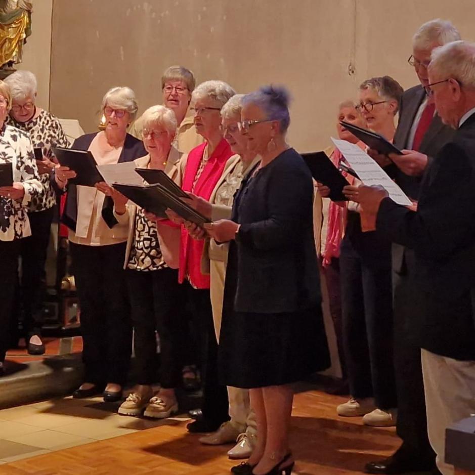 Das Foto zeigt den Kirchenchor Cäcilia Wanderath als Gruppe von etwa 15 bis 20 Personen in der Kirche. Die Gruppe steht in einem Halbkreis und hält Notenmappen in den Händen. Links im Bild steht der Chorleiter Klaus Bierbrauer, die die Gruppe dirigiert und mit erhobener Hand den Takt vorgibt. Die übrigen Personen stehen dicht beieinander auf einer leicht erhöhten Stufe und schauen in ihre Noten oder in Richtung der dirigierenden Person. Die Kleidung ist überwiegend festlich, vor allem dunkle Jacken, mit einigen helleren Oberteilen und farbigen Akzenten. Im Hintergrund ist der Innenraum einer Kirche zu sehen, mit hellen Wänden und einer Figur an der Wand. Der Boden besteht aus hellen Fliesen und Holz. Insgesamt zeigt das Bild eine Chorgemeinschaft.